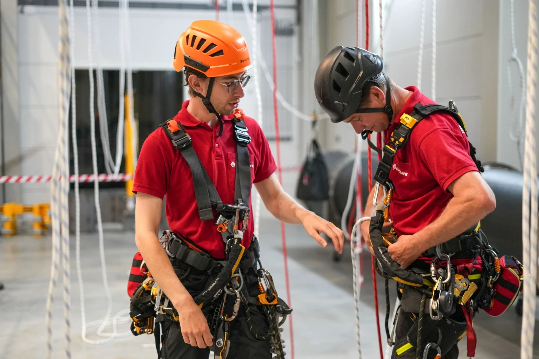 Twee personen die hun harnas goed trekken. Ze dragen een helm en staan in de trainingshal.