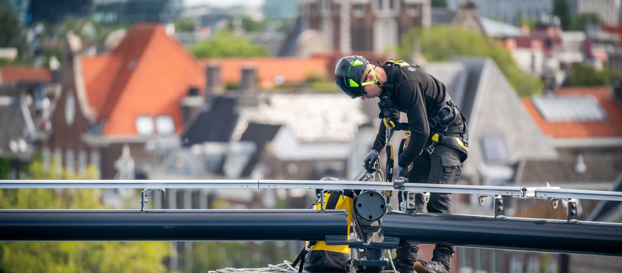 een man die op een dak staat naast een railsysteem, aangelijnd en met een rugzak op zak