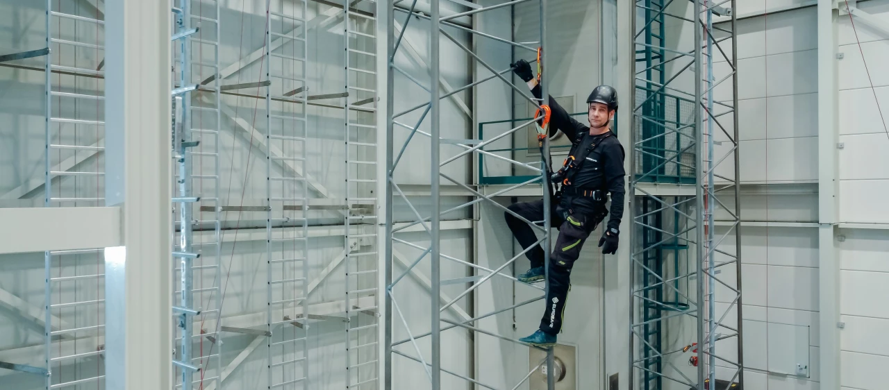 Een man die op een zendmast staat met een helm op en een harnas om