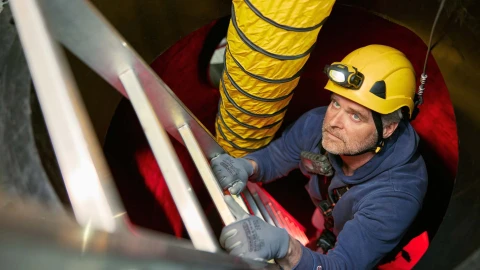 man klimmend op ladder met helm op en harnas aan in besloten ruimte